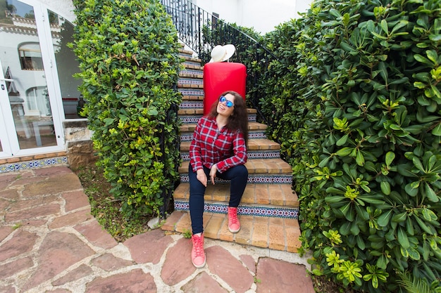 Travel, tourism and people concept - happy young woman sitting on stairs in sunny glasses with red suitcase and hat lying on it.