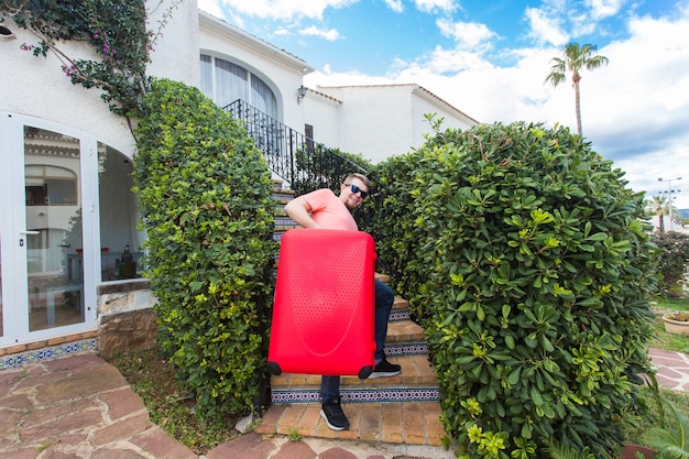 Travel, trip and holidays concept. young man with red suitcase in sunny glasses standing on stairs.