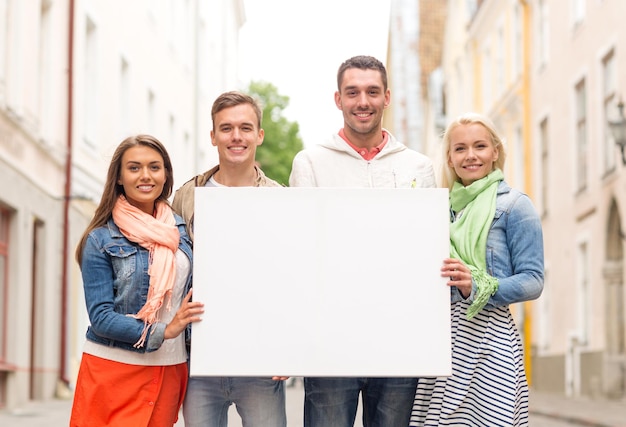 travel, vacation and advertising concept - group of smiling friends with blank white board in the city