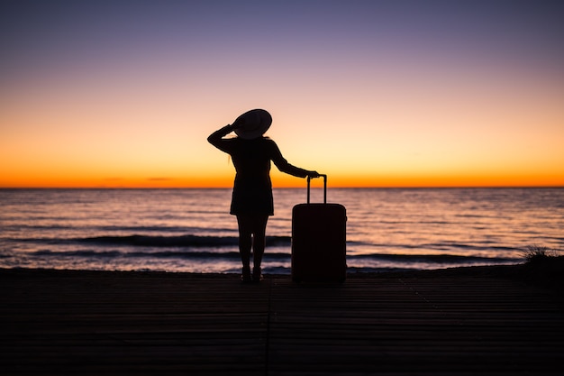 Photo travel, vacation and summer trip concept - silhouette of young woman in summer dress and hat looking to a sea.