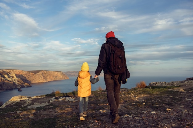 Traveler boy child with father walks on a cliff by the sea in jackets with his back to the sunset