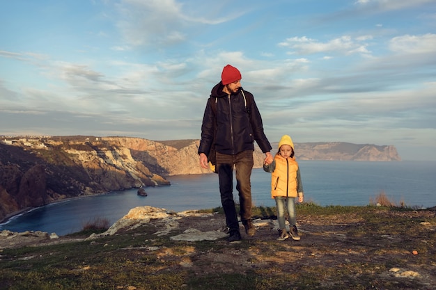 Traveler boy child with father walks on a cliff by the sea in jackets with his back to the sunset