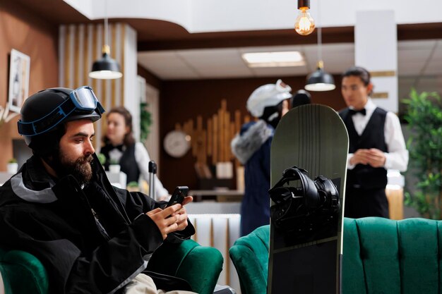 Traveler browsing on cellphone sitting on hotel reception sofa next to his snowboard. Male guest checking reservation on mobile device. Staff attending. Winter gear hints at ski resort vacation.