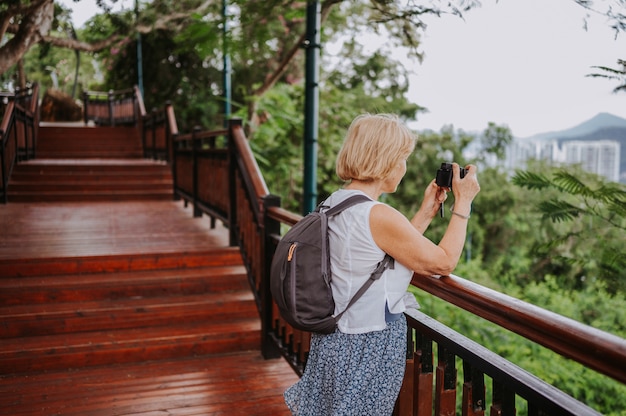 Traveler elderly senior backpacker woman  walking taking photos in tropical park, Travel adventure nature in China, Tourist beautiful destination Asia, Summer holiday vacation trip, Copy space