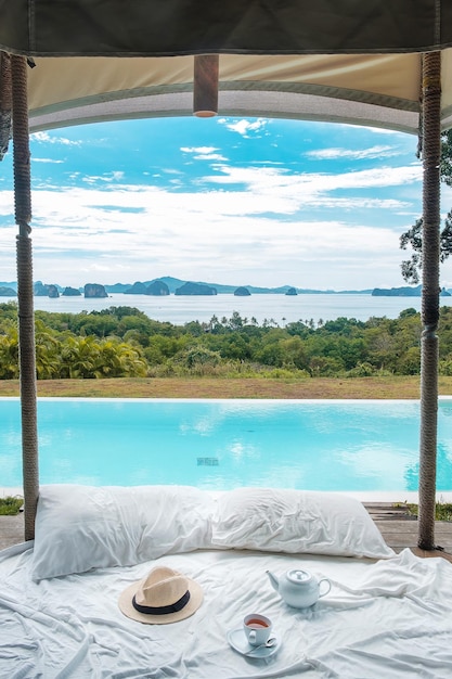 Traveler hat hot tea cup and teapot on white bed against Beautiful ocean view background Tourists relaxing in tropical resort with swimming pool travel summer vacation and freelance concept