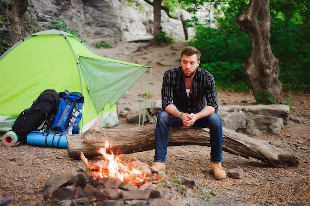 Traveler man relaxing, looking at the fire and dreaming at camping tent outdoors on nature