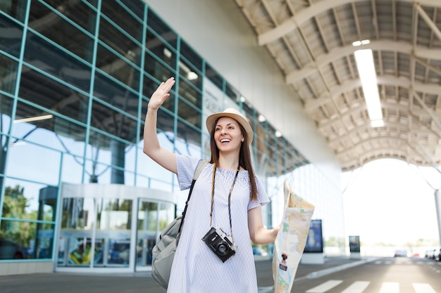 Traveler tourist woman with retro vintage photo camera, paper map waving hand for greeting, meeting friend and catch taxi at airport