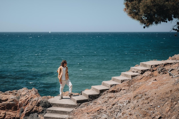 A traveler with long hair and bright closes stands on stairs with blue sea background