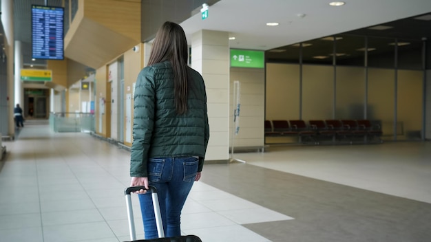 Traveler woman brunette is walking in modern airport terminal carrying suitcase back view Passenger is going to her flight in city airport