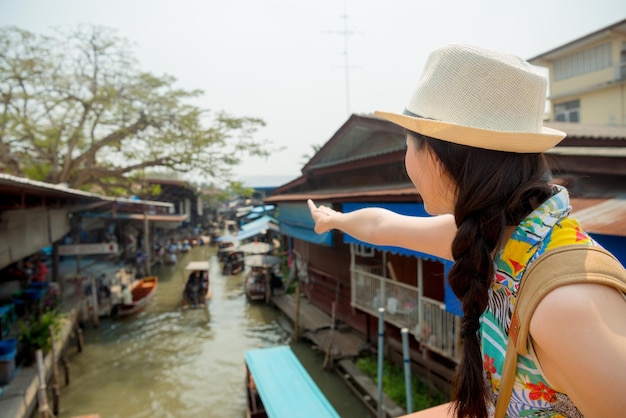 traveler young woman with backpack and cap view from back tourist traveler planning to take a boat going shopping and pointing hand for river in the Damnoen Saduak floating market, in Thailand, Asia.