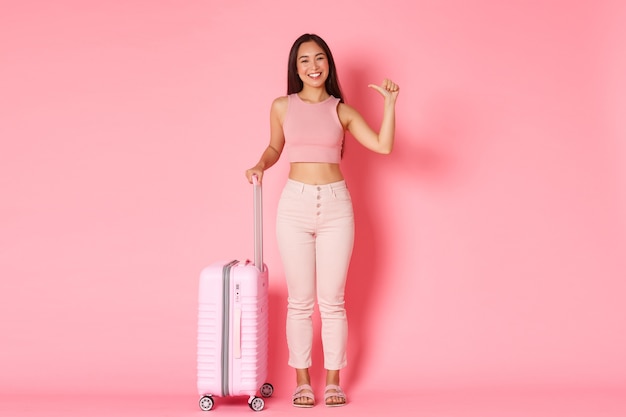 Traveling, holidays and vacation concept. Full-length of sassy and happy asian girl tourist standing over pink wall with suitcase and pointing at herself, explore countries