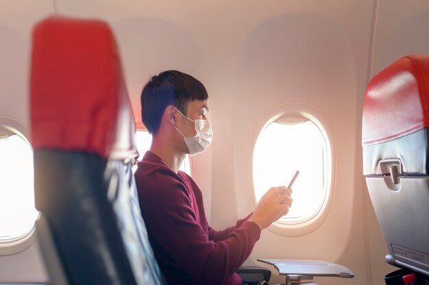 A traveling man wearing protective mask onboard in the aircraft using smartphone, travel under Covid-19 pandemic