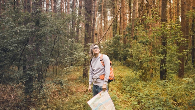 A traveling man with dreadlocks with a map in the forest A male traveler with a backpack and a map in his hand in the forest in cloudy weather