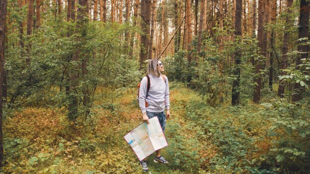 A traveling man with dreadlocks with a map in the forest A male traveler with a backpack and a map in his hand in the forest in cloudy weather