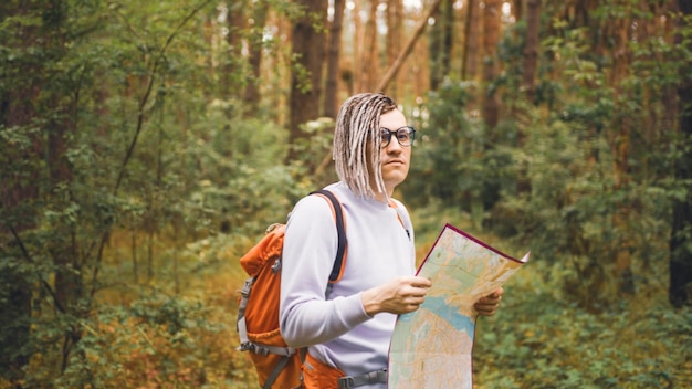 Traveling man with dreadlocks with map in woods Male traveler with backpack in woods reading map in cloudy weather