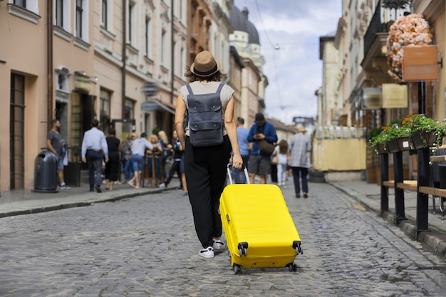 Traveling woman tourist in hat with backpack and suitcase walking along street of old tourist city, summer sunny day back view, walking people background