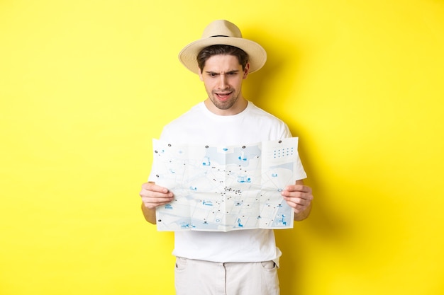 Photo travelling, vacation and tourism concept. man looking confused at map during trip, cant understand, standing over yellow background