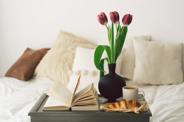 Tray with croissant, cup of coffee and tulip on white clean bed.