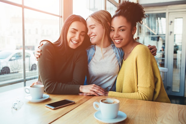 Tree young women are sitting together in a small cafe with big windows and embrace with each other.