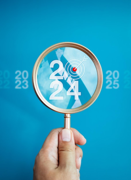 Trends searching business goals and Happy New Year 2024 concepts The big white 2024 year number with target icon inside the gold magnifying glass holding by hand on blue background vertical style