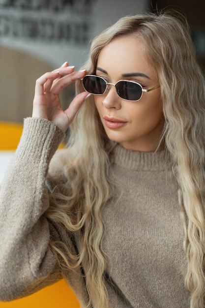 Trendy beautiful blondie girl in fashionable clothes with a beige sweater puts on vintage sunglasses and sits in a cafe near the window