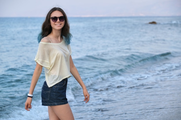 Trendy beautiful teenager girl with long colored blue hair in sunglasses and shorts walking along sea beach