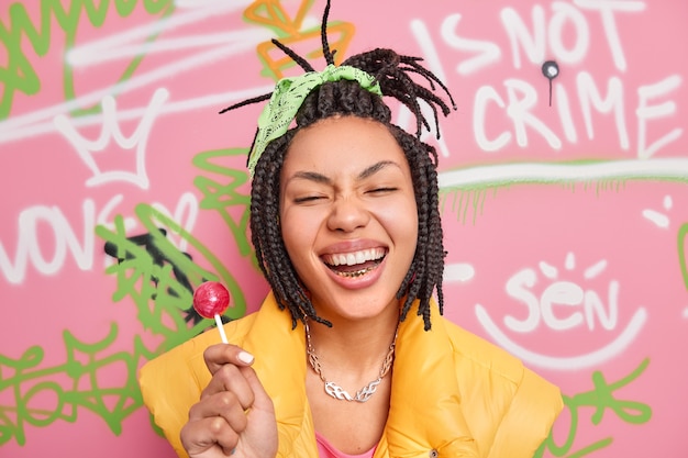 Trendy cheerful hipster girl smiles broadly holds lollipop has fun with teenagers of same age wears yellow vest poses against colorful graffiti wall