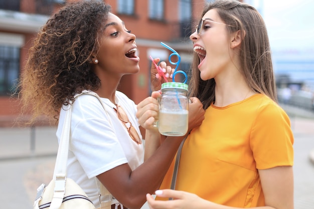 Trendy cool hipster girls, friends drink cocktail in urban city background.