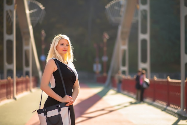 Trendy dressed blonde model with bright makeup posing at the bridge. Space for text