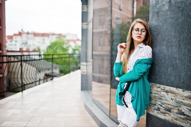 Trendy girl at glasses and ripped jeans against house on street.