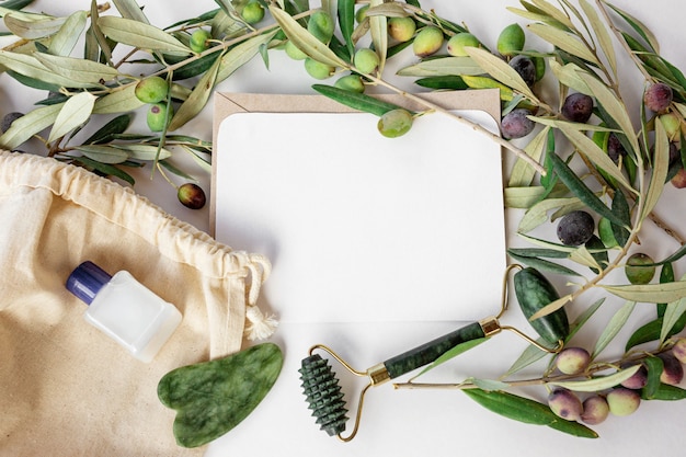 Trendy jade roller and gua sha green on natural white background with olive branches around, organic cotton bag, small white liquid bottle and copy space card. Flat lay, top view.