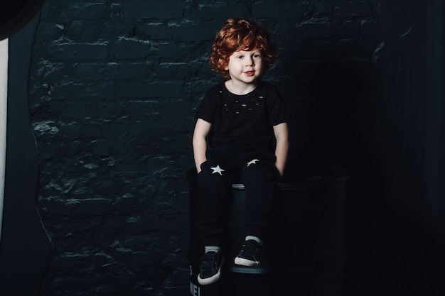 Trendy little boy in dark clothes posing in dimly lit room