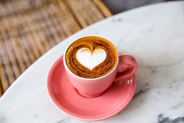 Trendy pink cup of hot cappuccino on marble table background. Heart shape latte art for symbol of love. One cup for morning routine.