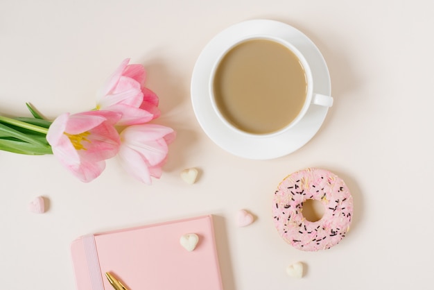 Trendy stylish concept for a female blogger, freelancer: pink diary, notebook, Donat, pink tulips and an cup of coffee  on beige background. Working from home place