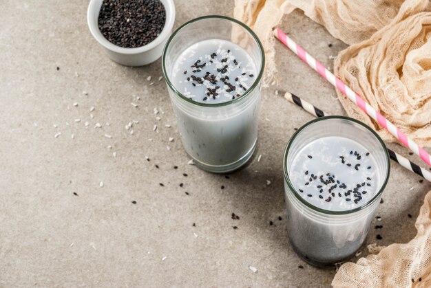 Photo trendy vegan food recipes, black sesame iced latte or smoothie with sesame seeds, dried coconut and almond milk, grey stone table,  copyspace top view