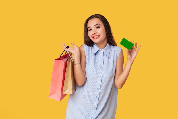 Trendy woman 20s in dress with long brown hair smiling while holding different shopping packages and card in hands isolated over yellow background