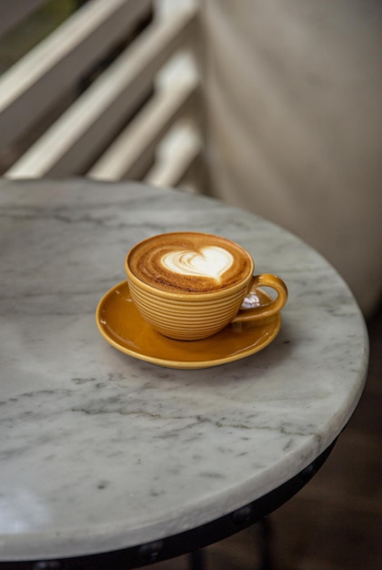 Trendy yellow cup of hot cappuccino on marble table background Heart shape latte art for symbol of love