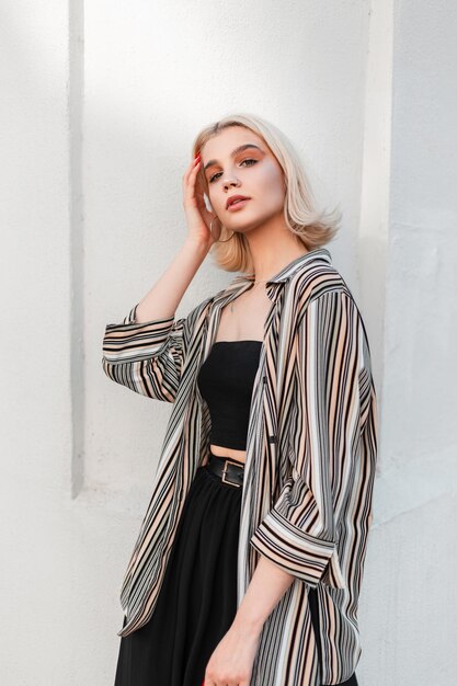 Trendy young beautiful woman with short blonde hairs in shirt black top and skirt stands near white wall