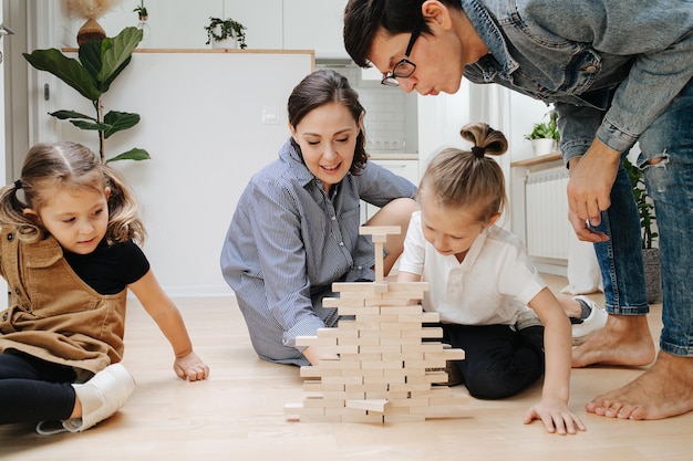 Tricky dad blowing on jenga wall while his son pulling a tile out of it Parents and children sitting on the floor