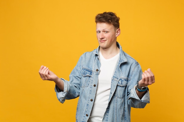 Tricky young man in denim casual clothes rubbing fingers showing cash gesture asking for money isolated on yellow orange wall background. People sincere emotions lifestyle concept. Mock up copy space.