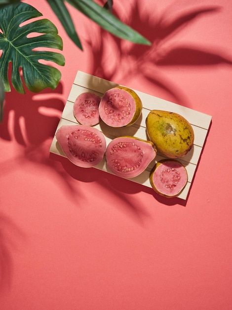 Tropical fruit concept Fresh whole and cut guava with pink pulp on a background of the same color under leaves with hard shadows