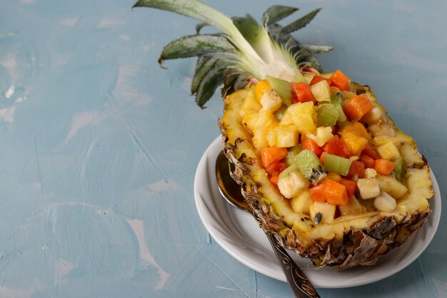 Photo tropical fruit salad in the half pineapple on white plate on a light blue surface, horizontal orientation, copy space, closeup