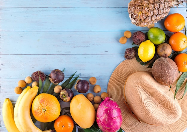 Tropical palm leaves, hat, many fruits on blue wooden background.