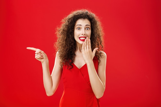 Troubled cute and tender silly woman with curly hairstyle in red stylish evening dress saying oops smiling with guilty look covering mouth with palm while pointing left over studio background.