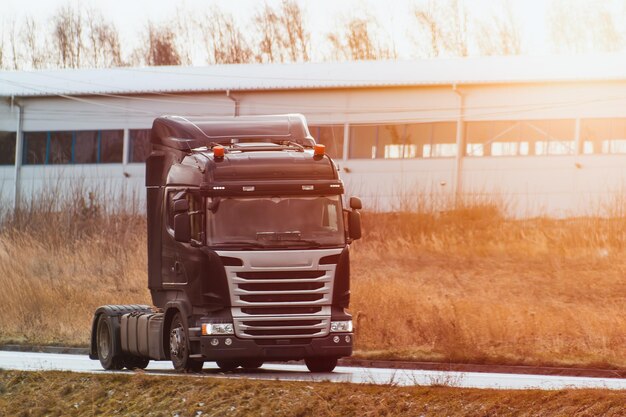 Photo a truck travels on an open road with no trailer attached european modern truck runs on diesel engine the road is flanked by wild grass and an industrial area in the distance