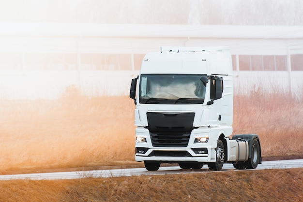 Photo a truck travels on an open road with no trailer attached european modern truck runs on diesel engine the road is flanked by wild grass and an industrial area in the distance