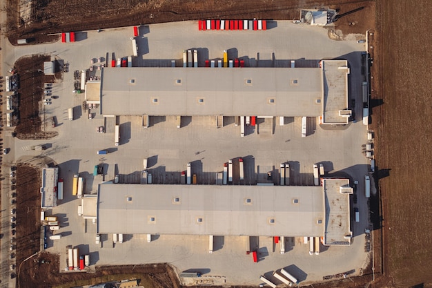 Trucks with trailers wait for the loading of goods for transportation in the loading warehouse. Aerial top view shot. A large transit cargo terminal with parking, trucks and trailers.