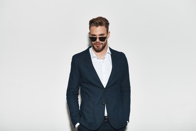 True elegance. Handsome young man in full suit keeping hands in pockets and looking at camera while standing against grey background