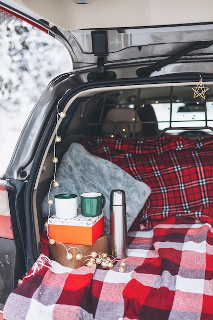 Trunk of car is decorated for celebration of Christmas and New Year in snowy winter forest Mug and thermos for drinking hot tea Cozy red plaid and gift box White picnic in woods