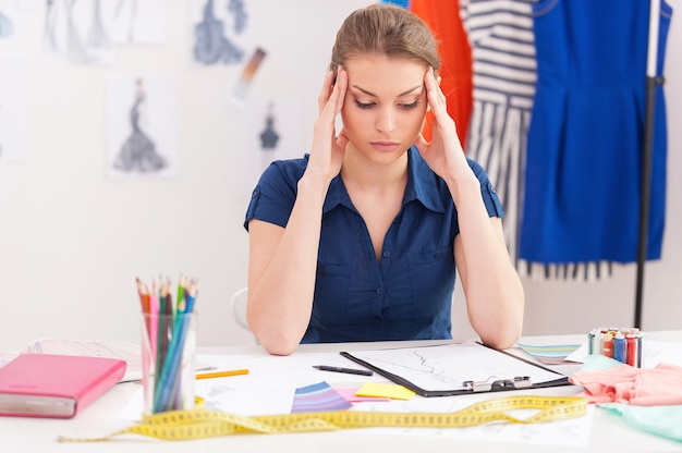 Trying to reach deadline. Depressed female fashion designer holding head in hands while sitting at her working place